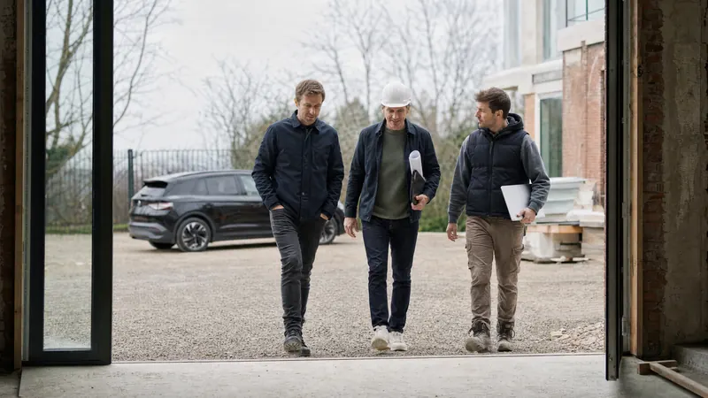 Drie mensen met plannen en een bouwhelm komen een gebouw in verbouwing uit lopen, de elektrische wagen staat achter hen geparkeerd