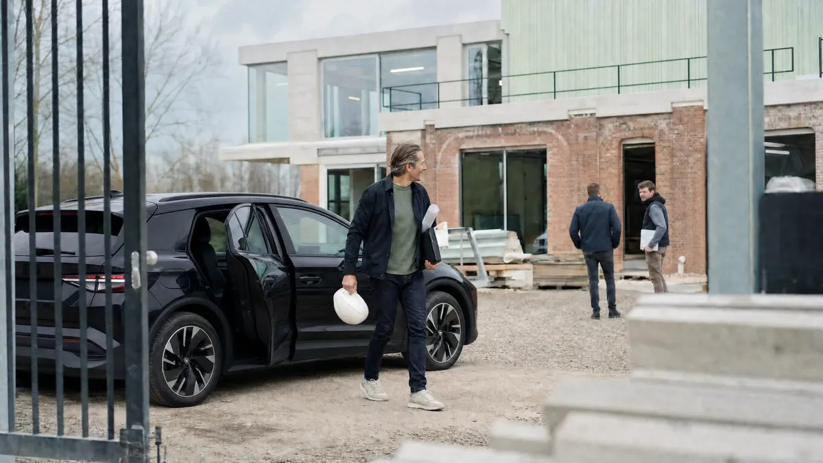 Projectleider met een witte bouwhelm onder de arm stapt uit een zwarte elektrische wagen, voor een gebouw in verbouwing