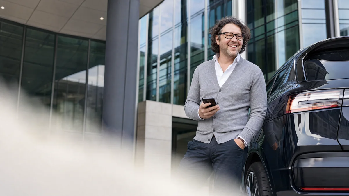 Een man met bril en grijs vest staat glimlachend met zijn telefoon in de hand naast de achterkant van een zwarte elektrische SUV, voor een modern glazen kantoorgebouw, gezien vanaf straatniveau