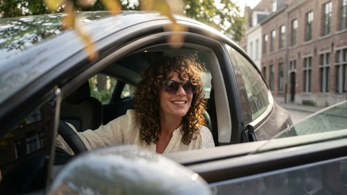 Een vrouw met krullen lacht achter het stuur van een elektrische wagen in een Vlaamse straat