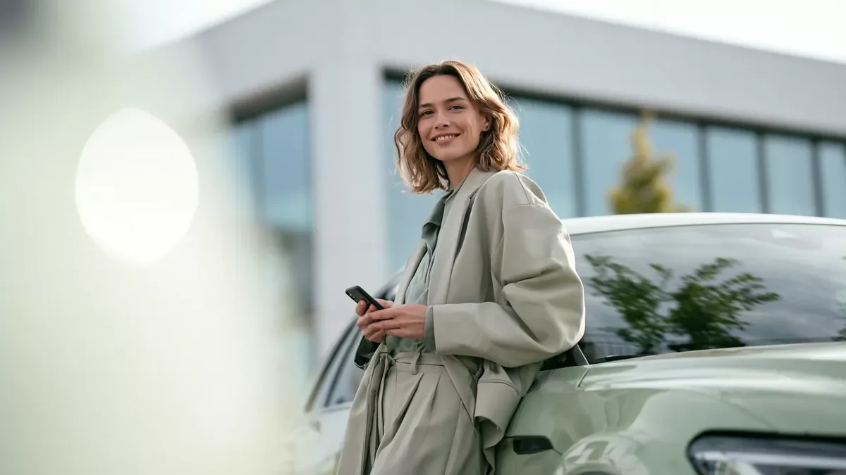 Een vrouw met haar telefoon in de hand leunt tegen een elektrische wagen bij een kantoorgebouw