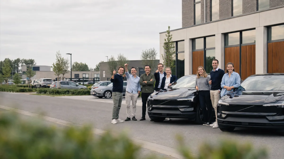 Een team van zeven collega's staat lachend naast twee zwarte elektrische Volvo's voor een modern kantoorgebouw op een bedrijvenpark